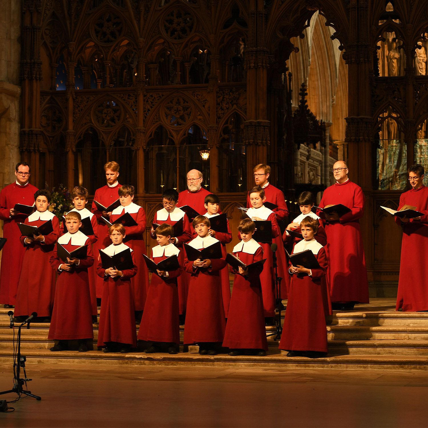 The Choir stand in front of a wooden screen at Winchester Cathedral, wearing red cassocks