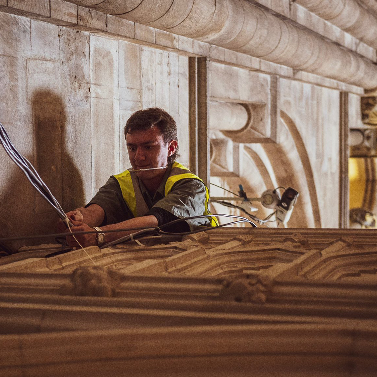 An engineer wearing a yellow fluorescent vest leans over a stone screen, holding cables and wires.