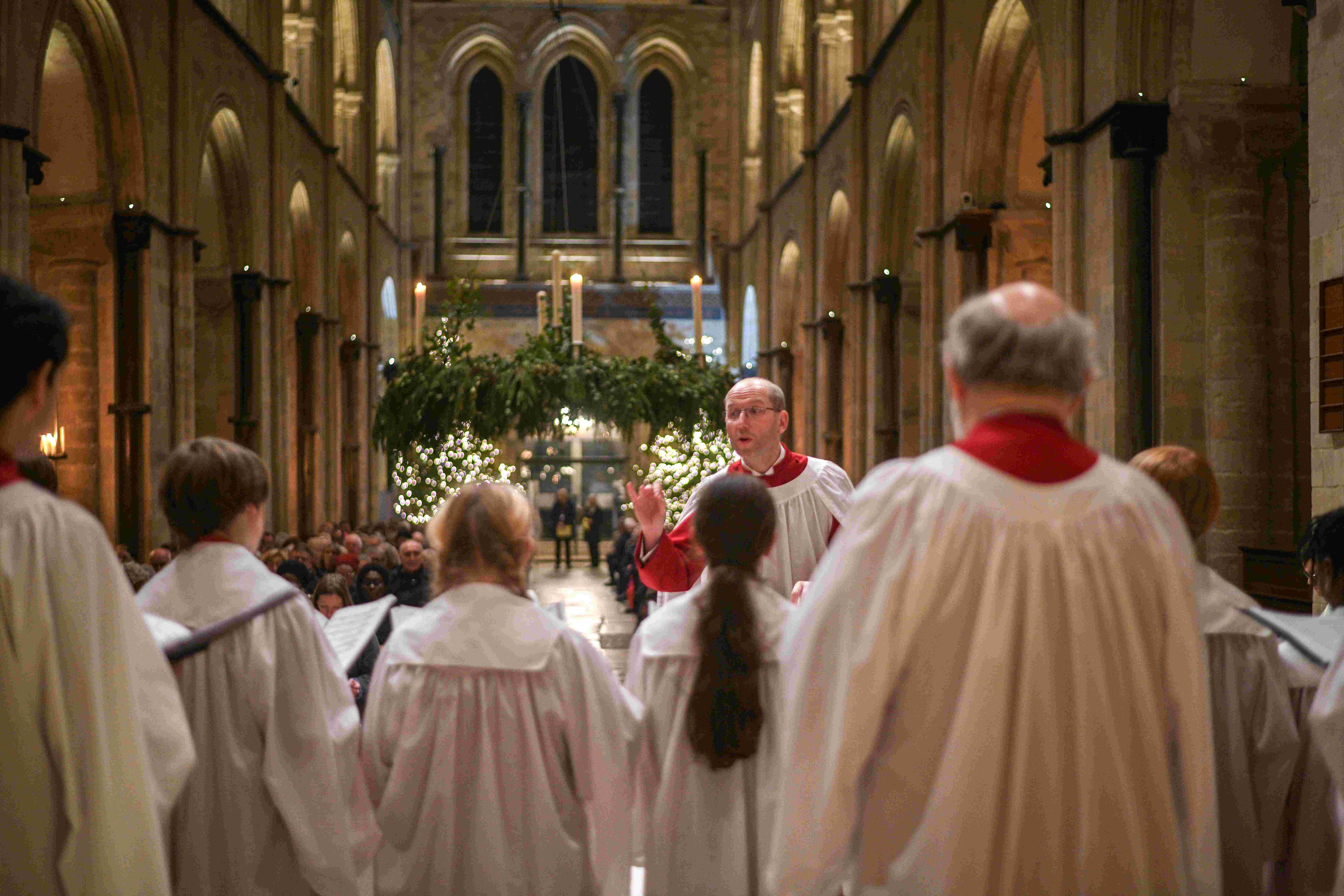 In the foreground are adult and child choristers in white robs, mid ground is a man in white choral robes conducting,  in the background are people sitting in seats and above them Christmas foliage