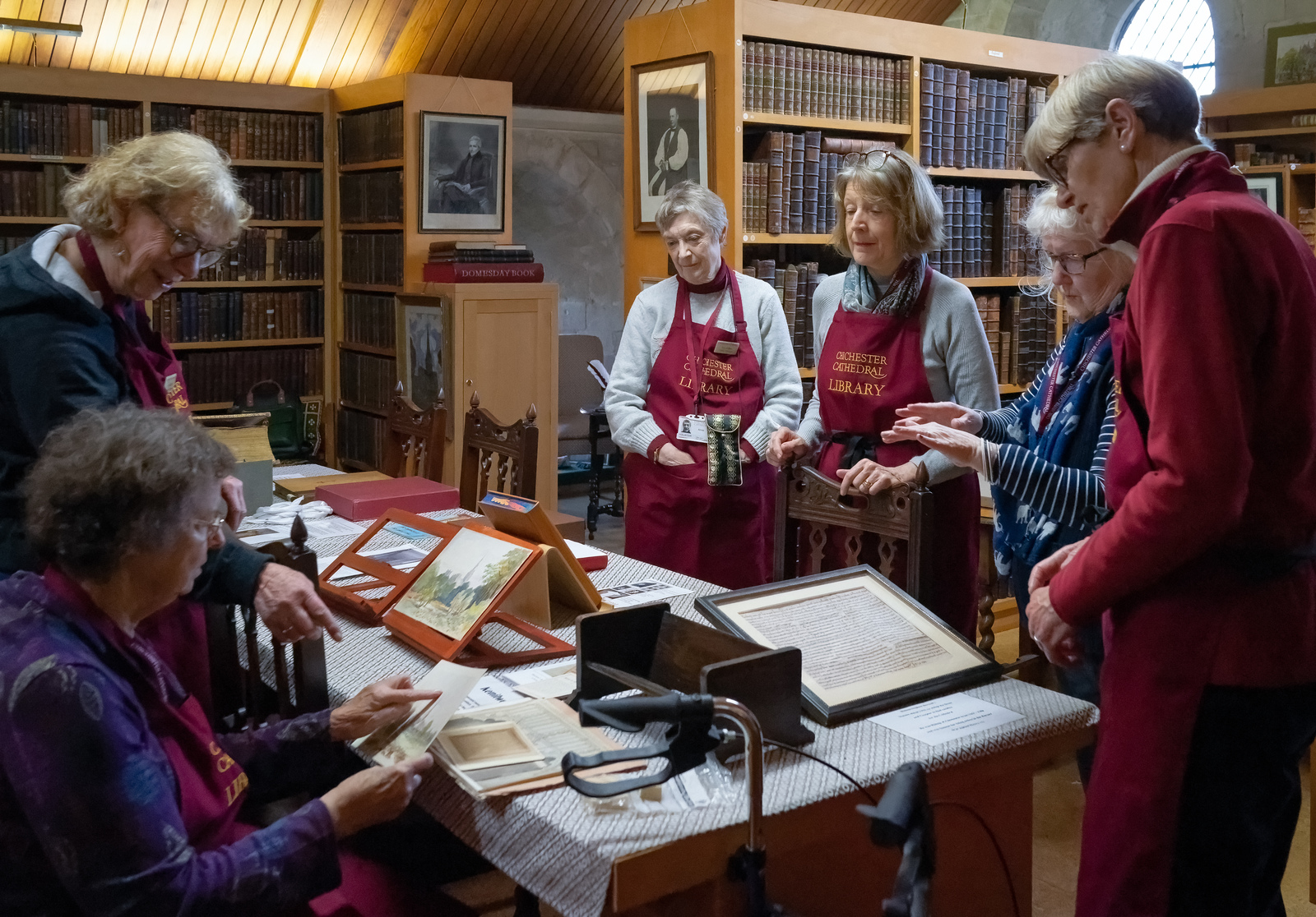 Six women standing (some wearing Chichester Cathedral Library Aprons)around a table looking at historic artefacts, bookshelves in the background.