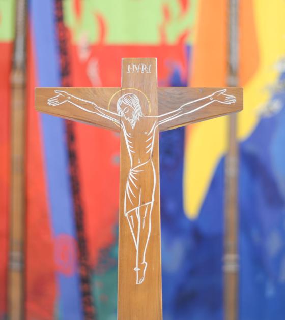 A wooden crucifix sits in front of a multi-coloured tapestry, with splashes of red, blue, green and yellow