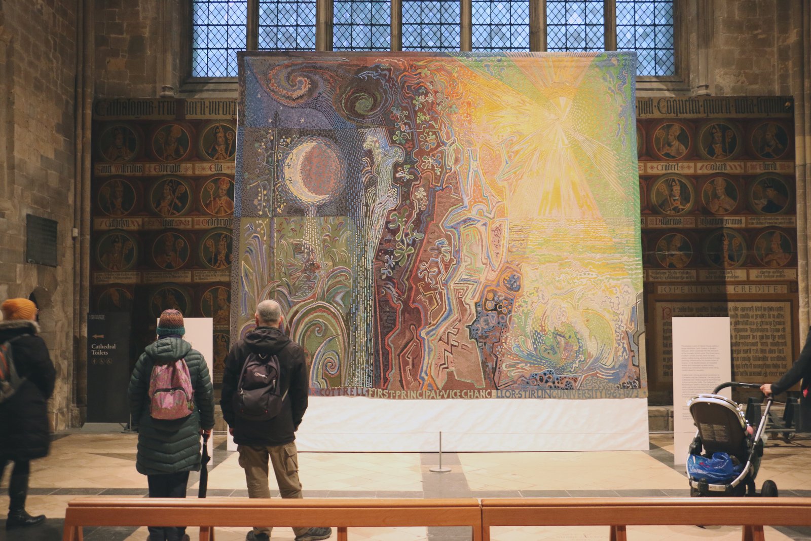 People admiring the large tapestry with their backs to the camera 