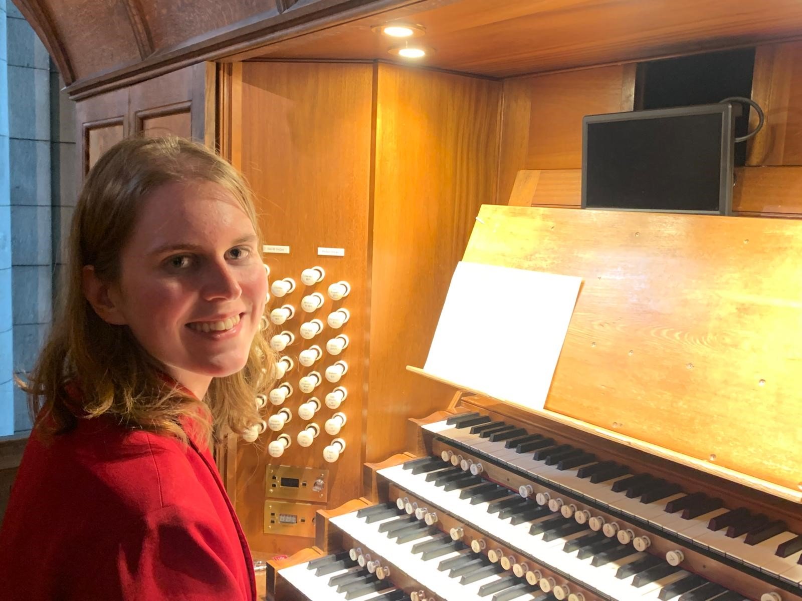 A young woman in her early 20s, with blonde-brown hair, wearing a red jacket, sits at an organ console, facing the camera and smiling warmly.